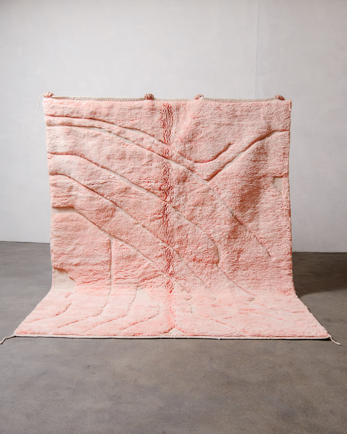 Berberlin Beni M'rirt Rug made of heavy wool pile in coral pink and cream being held up in front of a white wall and concrete floor.